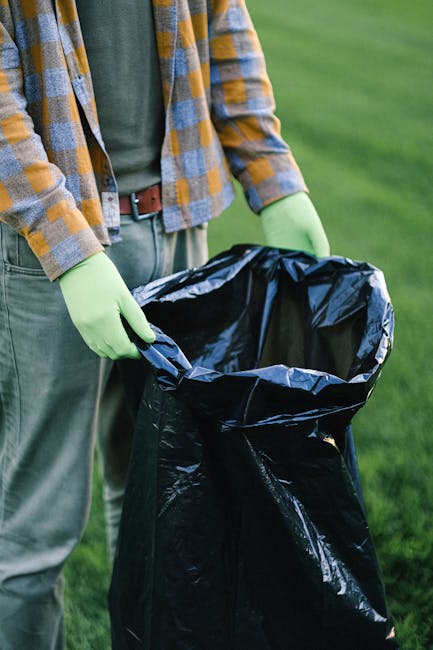 A waste collection operative, wearing a red and yellow high-visibility uniform, operates a large, red, industrial rubbish truck on a roadside. The truck has a rectangular rear loading section with mec