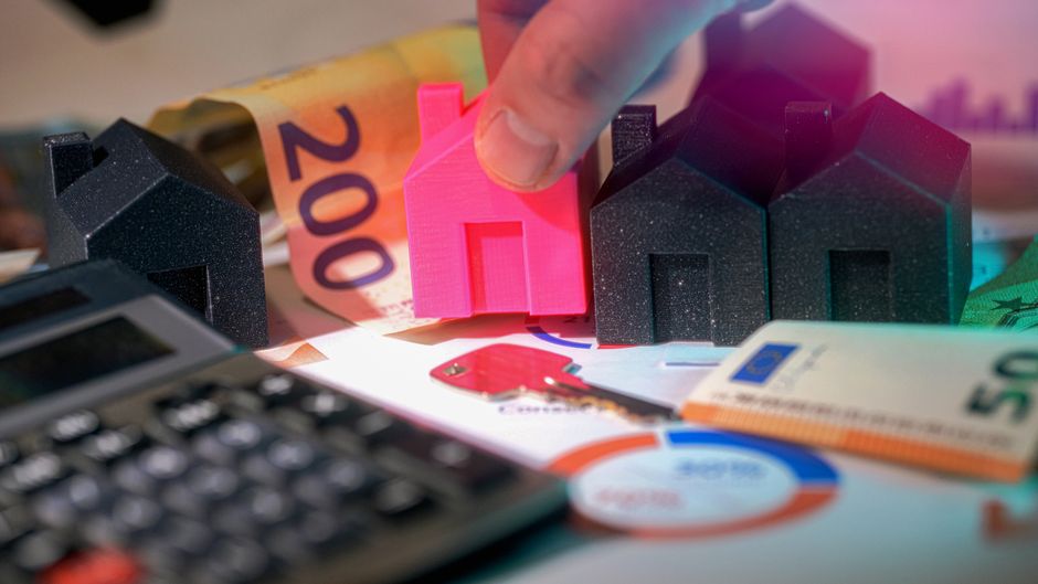 A close-up view of a person's hand placing a small, bright pink plastic house model among three additional house-shaped objects made of black foam material, positioned on a surface scattered with vari